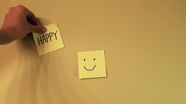 Human hand placing a yellow sticky note with the word happy next to another note with a smiling face drawn on it, representing joy, positivity, and sharing good feelings