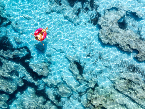 Aerial drone view of woman relaxing on pink inflatable ring in clear turquoise sea above rocks. Summer vacation, Mallorca, Balearic Islands, Spain. Top down view of swimming girl, blue water. Travel