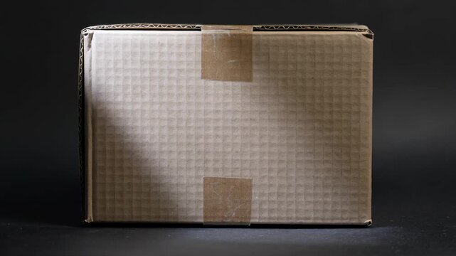 A plain cardboard box with tape on a dark background. The box is closed and has a textured surface, suitable for shipping or storage purposes.