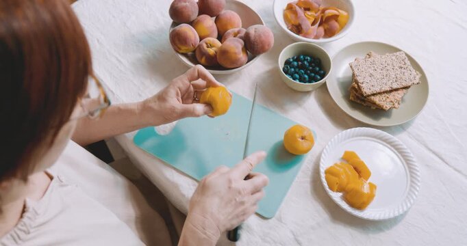 Woman cutting the peach for make sweet sandwiches crispbread with cream cheese, blueberry, slices of peach, coconut flakes and honey for summer picnic or brunch