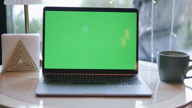Laptop with green screen sits on white table. Coffee mug rests beside laptop. Natural light shines through window. Decorative triangular object placed near laptop