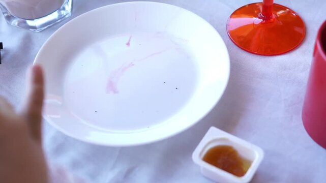 A young kid uses a knife to spread sweet apricot or peach jam over a slice of whole grain bread on a white plate during breakfast