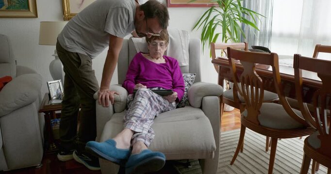 A moment between mother and son: a son leans in to whisper something to his elderly mother, who is engrossed in her tablet whilst sitting comfortably in an armchair.