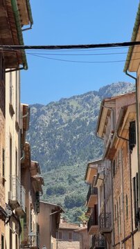 Charming street in Soller, Mallorca, framed by traditional stone buildings and lush greenery, with the dramatic Tramuntana mountains rising in the background.