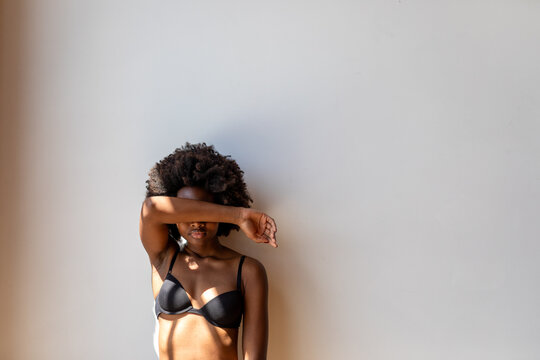 Person in black lingerie stands against a white wall, arm covering face.