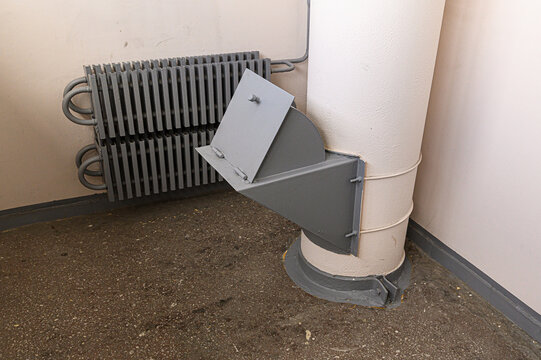 grey metal garbage chute with an open hinged flap is mounted in corner; adjacent, dark grey ribbed radiator is fixed to beige wall above speckled concrete floor