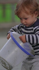 Fototapeta premium Young child examining colorful toy bucket at public playground during outdoor activity highlighting curiosity, sensory discovery and playful early childhood learning