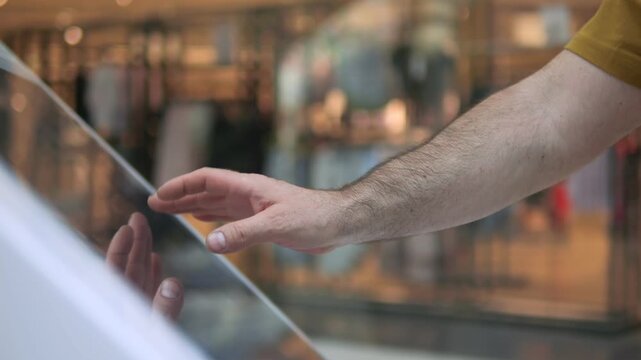Hand interacts with glass display in retail store. Fingers trace surface gently. Background shows blurred clothing racks. Scene captures modern shopping experience. Focus remains on hand motion