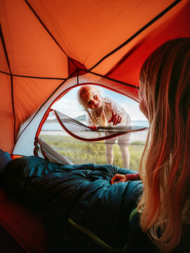 Family travel camping summer vacation - child girl peeking into tent entrance to wake her mother in the morning, parent woman laying in her sleeping bag gear, happy candid emotions adventure lifestyle