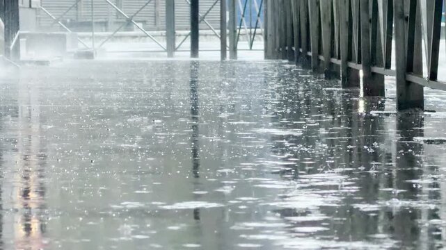 Heavy raindrops strike the flooded sidewalk to create a loud, rhythmic splashing sound. The water pools on the concrete, resulting in a continuous watery roar.
