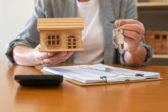 Real estate agent at desk holding wooden house model and keys over mortgage documents with pen and calculator, symbolizing home purchase, financing and property investment planning