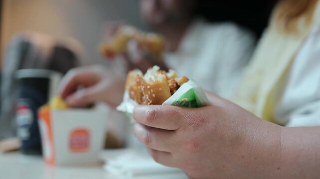 Detailed view of male or female hands holding a tasty fried chicken sandwich wrapped in green paper, eating fast food indoors.