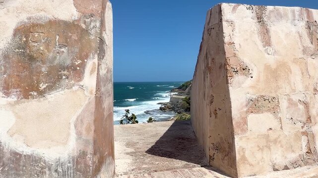 View between weathered stone walls of a historical fort, revealing the rugged Caribbean coastline, crashing ocean waves, and a traditional sentry box in San Juan, Puerto Rico.