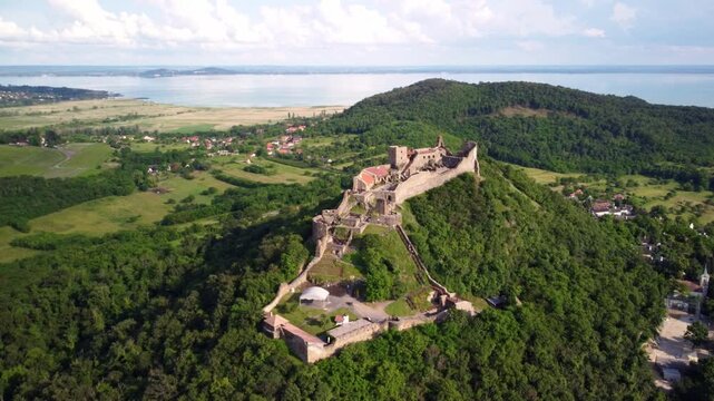 Szigliget Castle near Lake Balaton in Hungary during a drone orbit above the forested hill with views of the lake and surrounding landscape.