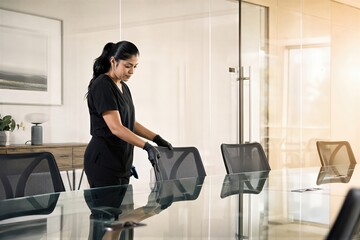 Fototapeta premium Hispanic woman cleaner sanitizing office chair with spray. Professional janitor cleaning glass meeting room table surface. Workplace maintenance and hygiene service concept.