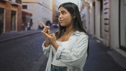 Fototapeta premium Young hispanic woman pointing with index finger and counting gesture on narrow cobblestone street; uncertainty and deliberation.