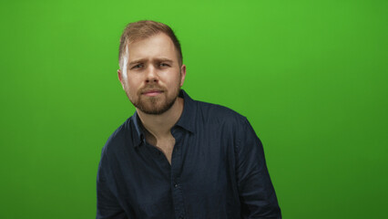 Naklejka premium Man tilts head, chest exposed and wearing dark blue shirt in studio with bright green backdrop; thoughtful introspection.