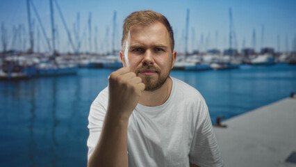 Fototapeta premium Man with clenched fist at port pier wearing white tshirt with boats in harbor; determination resilience strength.