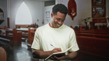 Fototapeta premium Man writing in notebook with pen in a church, standing by pews and smiling while holding book; reflection serenity.