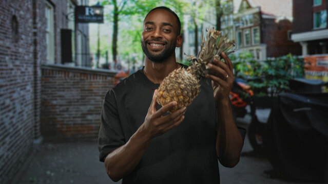 Man smiling while holding a pineapple with bare hands on a city street by a brick building and planters; casual joy summer.