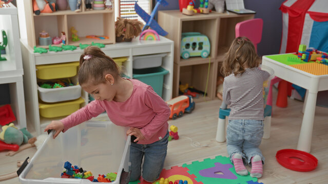 Girl kneels, hands in clear plastic toy box sorting colorful blocks on foam mat inside a playroom building; playful cooperation.