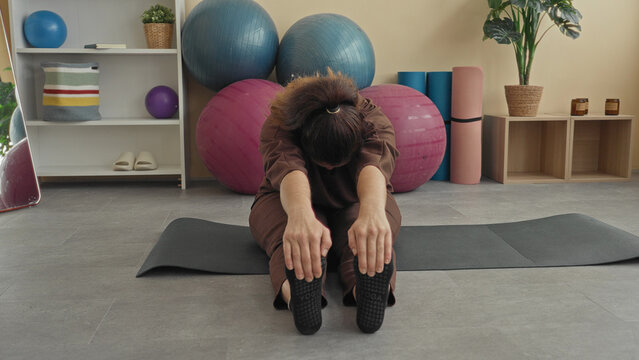 Woman on yoga mat reaches hands to toes while seated, stretching hamstrings in a fitness studio with exercise balls and foam rollers visible; calm focus.