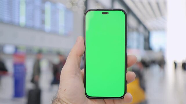 Hand holds phone with green screen at busy station. Background shows travelers and digital boards. Ideal for app demos, travel ads, or digital interface concepts