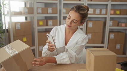 Woman holding parcel and smartphone while seated at table with stacked boxes and shelving in...