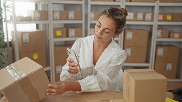 Woman holding parcel and smartphone while seated at table with stacked boxes and shelving in building shipping area; small business focus.
