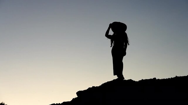 Mystical silhouette of a young woman taking off her sun hat on top of a rocky hill, then sitting down in shadow against a twilight sky