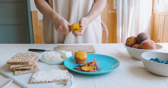 Anonymous woman peeling the peach for make sweet sandwiches crispbread with cream cheese, blueberry, slices of peach, coconut flakes and honey for summer picnic or brunch