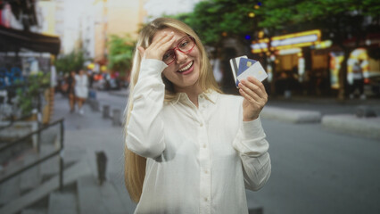 Obraz premium Woman holding a bunch of credit cards, hand to forehead gesture and smiling on a busy city street with shop lights and pavement; relief savings.
