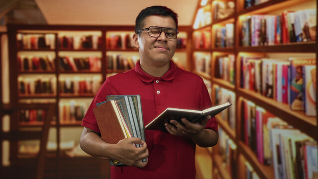 Young man reading an open book while holding stacked books in a library aisle; concentration learning ambition.