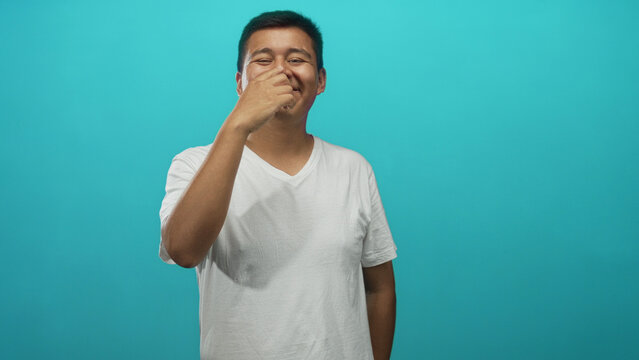 Young hispanic man pinches nose in turquoise studio wearing white t shirt and smiling at camera; disgust reaction.