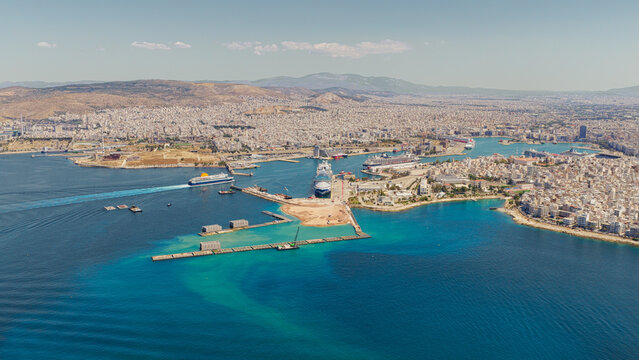 Aerial drone view of Piraeus port in Athens, Greece on a sunny day. A ferry departs near a docked cruise ship, surrounded by vibrant blue waters and the sprawling urban cityscape.