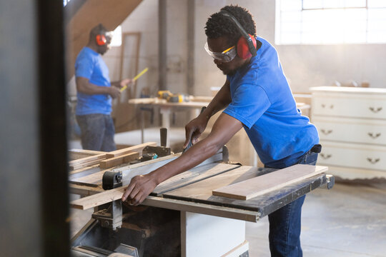 African American male coworkers guiding wooden plank through table saw in workshop, copy space