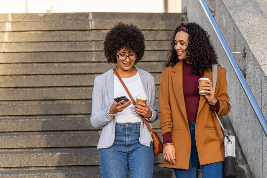 Two African American women wearing blazers walking down wide concrete stairs holding coffee cups