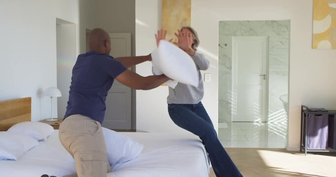Diverse couple exchanging white pillow swings in bedroom, man initiating hit, woman falling for fun