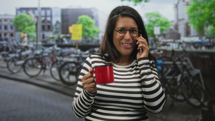 Fototapeta premium Woman holding a red mug and phone to her ear, smiling by rows of parked bicycles on a street; warmth joy connection.