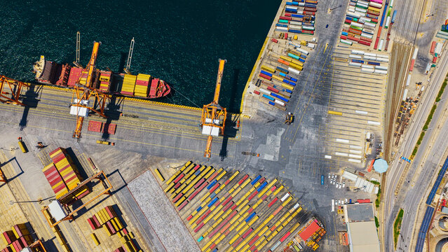 Aerial view of a global logistics hub with a massive container ship being loaded by cranes at a busy port terminal amid rows of colorful shipping containers and quay activity