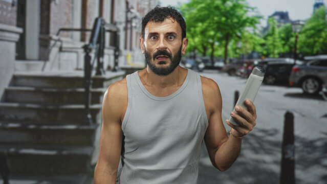 Man with beard in tank top holding shampoo bottle in his hand on an urban street stoop near parked cars, expression puzzled; grooming uncertainty.