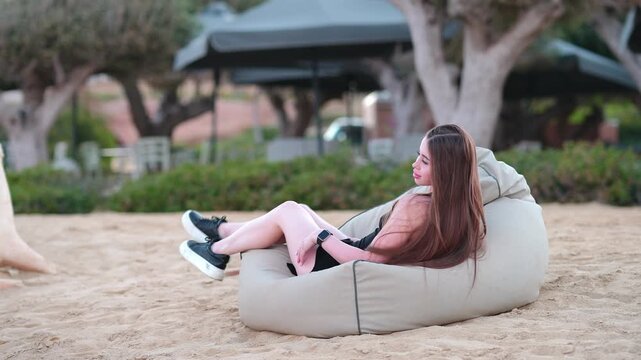 Beautiful young woman with long hair sitting on a comfortable beanbag chair. She is relaxing on a sandy beach, enjoying the leisure time