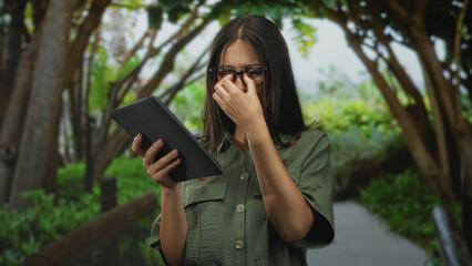 Woman pinching nose and holding tablet on forest path under trees, wearing glasses and green shirt  disgust. © Krakenimages.com