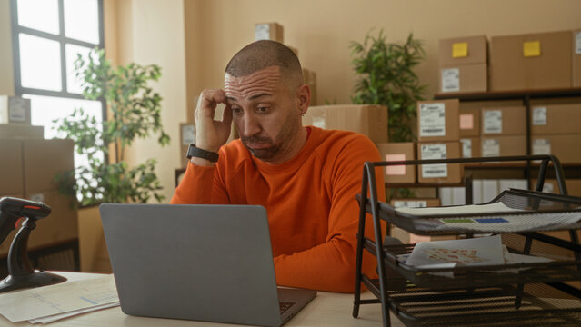 Man in orange sweater checks laptop and watch, hand on chin, barcode scanner on desk in building; small business frustration.
