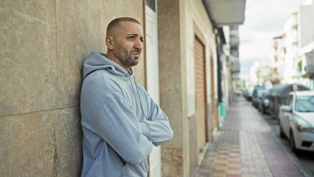 Man leaning against a building with arms crossed, face turned toward the street, wearing a light hoodie and small earring; quiet contemplation.