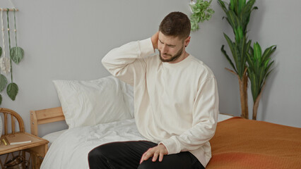 Fototapeta premium Man sitting on the edge of a bed yawning while stretching and rubbing his neck in a studio; morning routine sleepy.