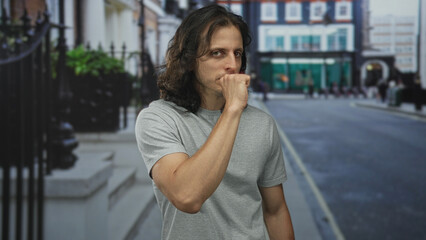 Man pressing hand to mouth coughing on a busy city street, wearing grey tshirt and long dark hair  quiet concern. © Krakenimages.com