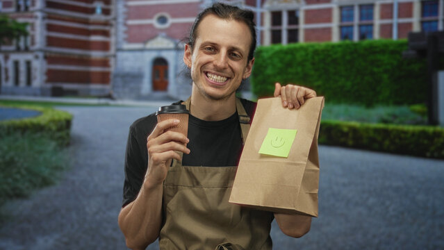 Man in apron holds a takeaway coffee cup in his right hand and a paper bag with a sticky smiley note while smiling in front of a brick building; friendly takeaway happiness.