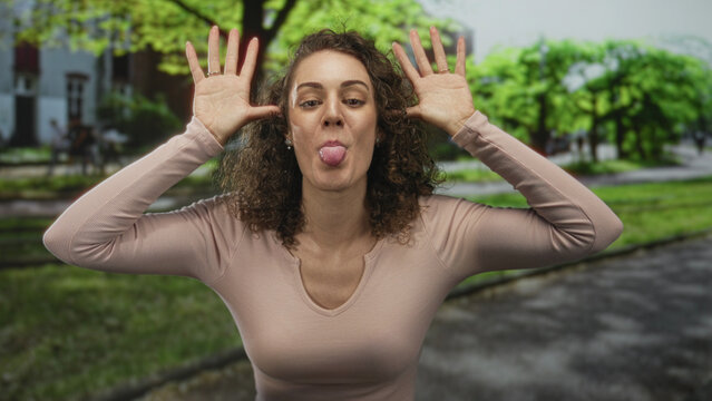 Young hispanic woman sticks out tongue and lifts hands to ears in park near green trees and paved walkway; playful mischief.