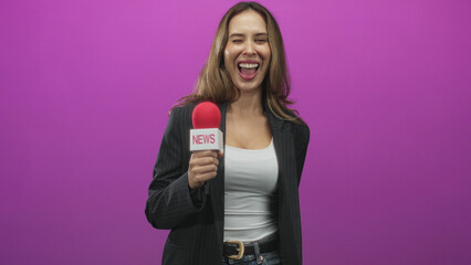 Fototapeta premium Woman reporter holding news microphone and smiling, points finger at camera in studio with pink backdrop, wearing black blazer and white tank; joyful confidence.
